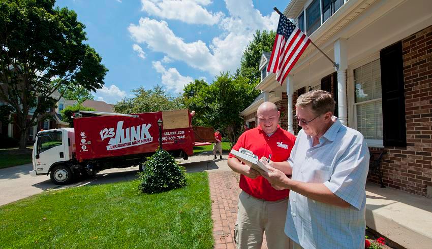 Junk removal worker in red shirt holding a clipboard and talking with a homeowner outside a house with a 123JUNK truck in the background