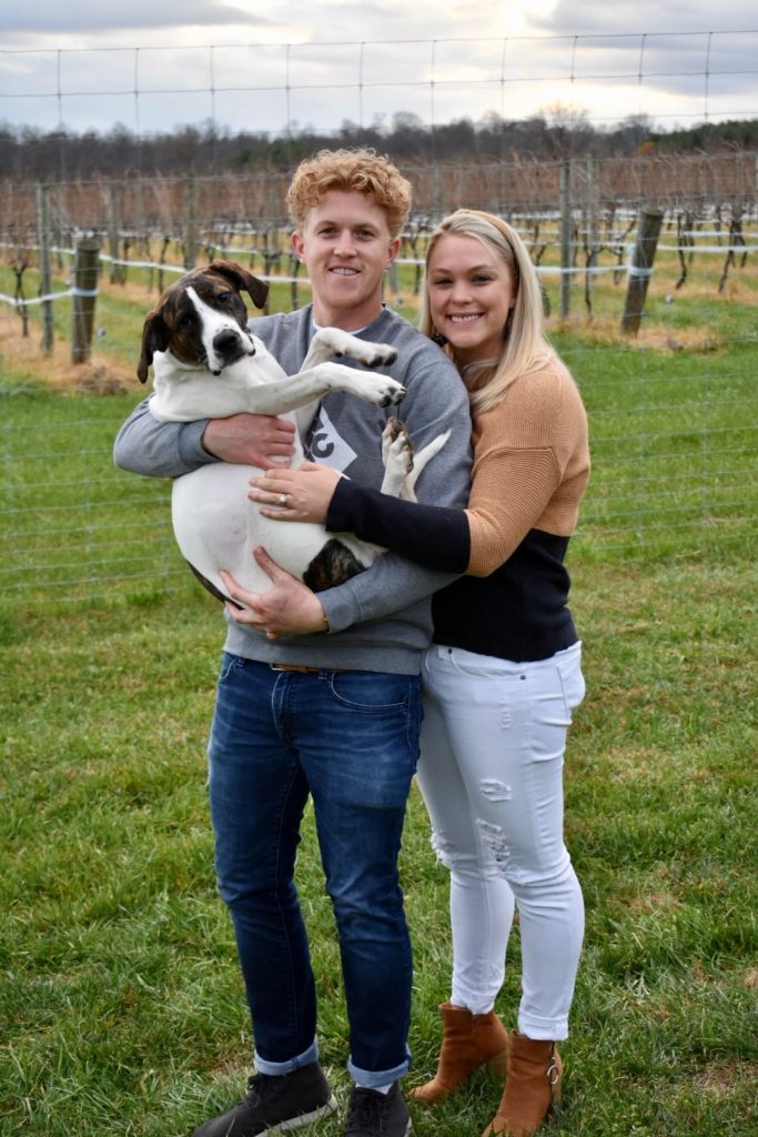 Smiling man and woman standing together in a field, with the man holding a large dog in his arms