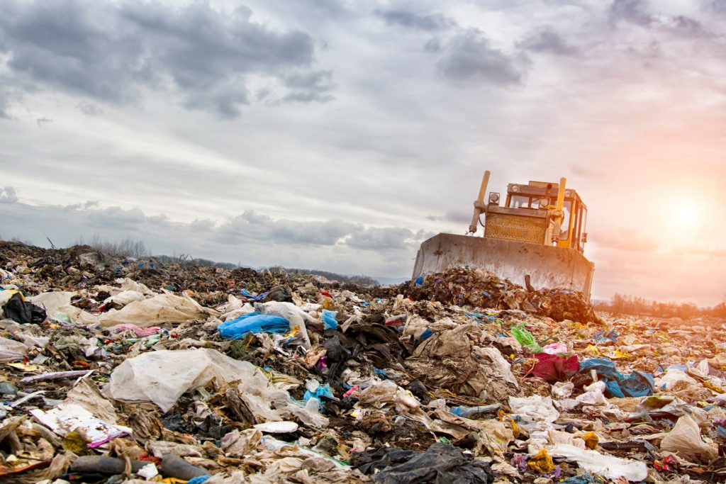 Landfill scene with a bulldozer pushing piles of mixed trash under a cloudy sky with sunlight breaking through