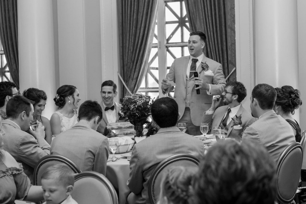 Black and white photo of a man giving a speech with a microphone at a wedding reception while guests, including the bride and groom, laugh and smile