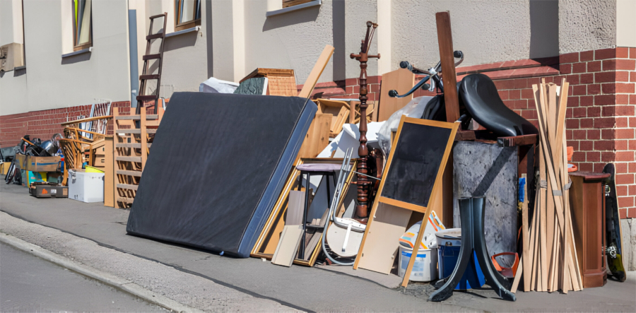 Large pile of discarded furniture, mattresses, and household items stacked along a sidewalk beside a brick building