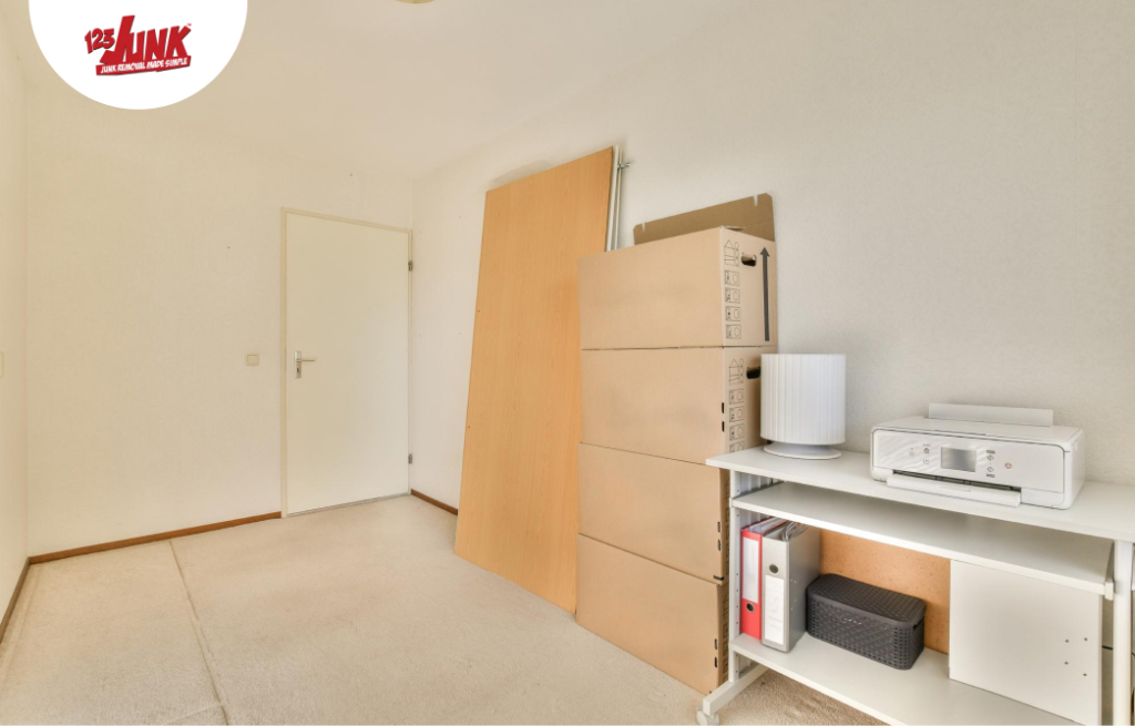Empty room with stacked cardboard boxes, a disassembled table, and a small white shelf holding a printer and binders