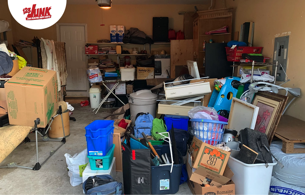 Cluttered garage filled with boxes, bins, furniture, and miscellaneous household items stacked to the front